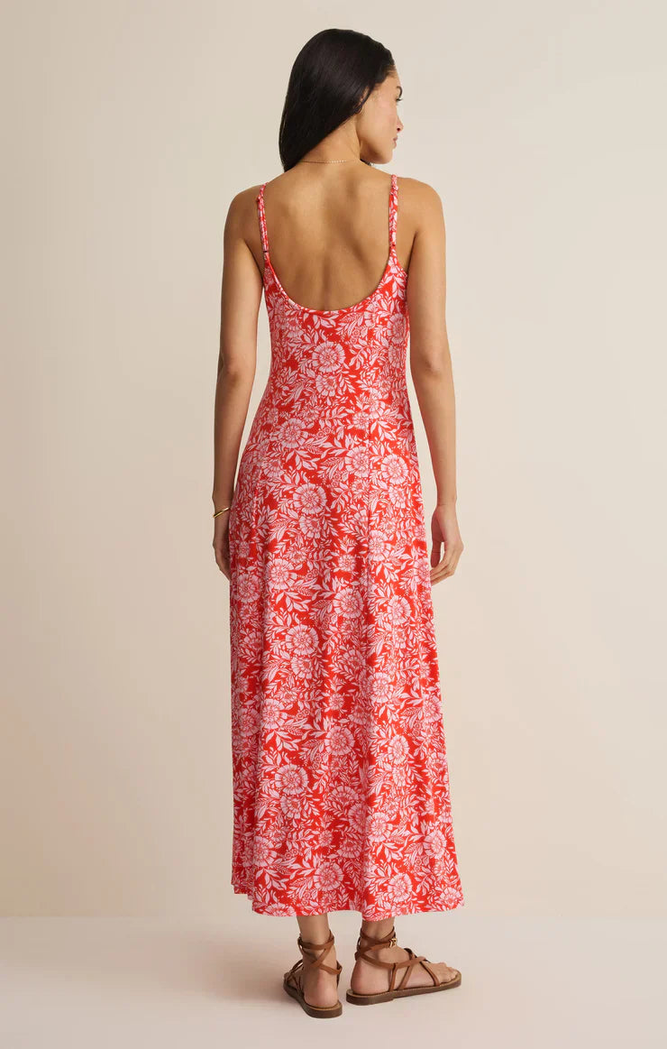 A woman faces away, wearing the Zertz CHARISMA ONA FLORAL MIDI DRESS with adjustable spaghetti straps, a sleeveless red and white floral design, styled with flat brown sandals, set against a plain beige background.