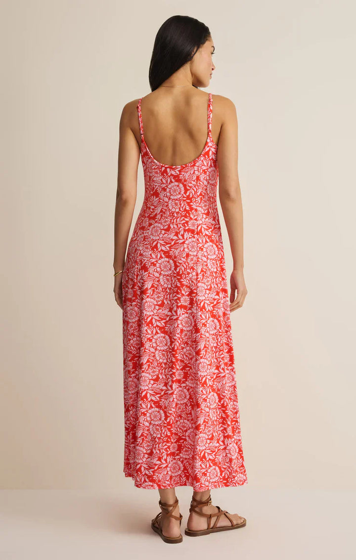 A woman faces away, wearing the Zertz CHARISMA ONA FLORAL MIDI DRESS with adjustable spaghetti straps, a sleeveless red and white floral design, styled with flat brown sandals, set against a plain beige background.
