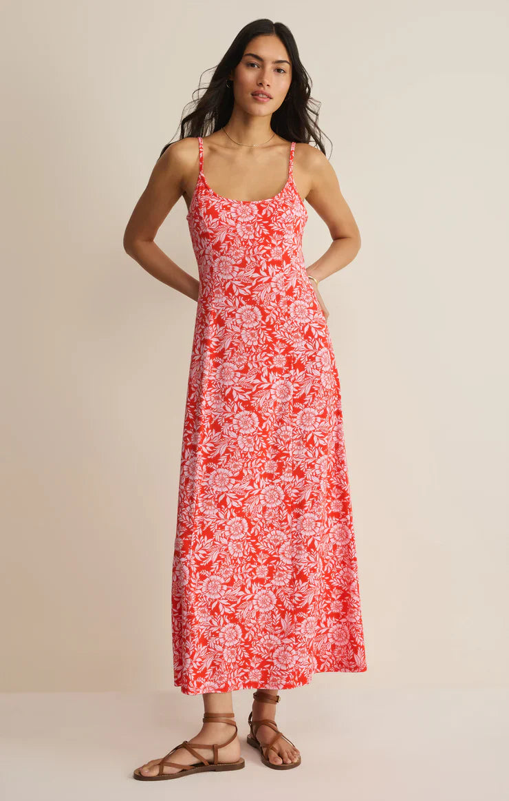 A woman with long dark hair stands against a plain background wearing the Zertz CHARISMA ONA FLORAL MIDI DRESS, featuring adjustable spaghetti straps and a red and white floral print, paired with brown strappy sandals and her hand on her hip.