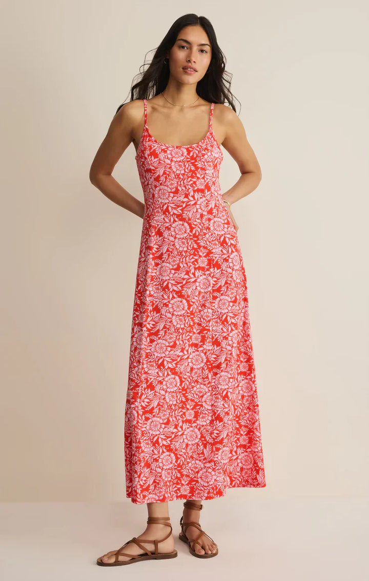 A woman with long dark hair stands against a plain background wearing the Zertz CHARISMA ONA FLORAL MIDI DRESS, featuring adjustable spaghetti straps and a red and white floral print, paired with brown strappy sandals and her hand on her hip.