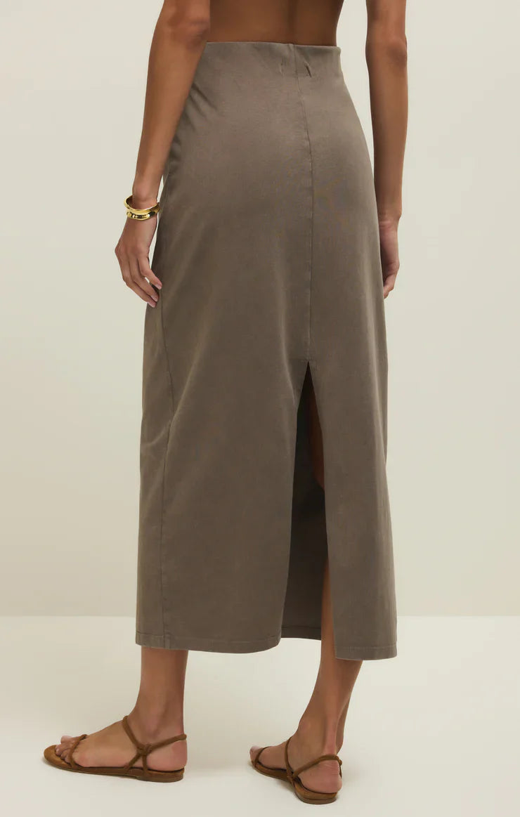 A person wears the Zertz GAIL MIDI SKIRT PERFECTLY TAUPE, a high-waisted cotton jersey midi skirt with an elastic waistband and back slit, styled with brown strappy sandals and a gold bracelet against a plain, light background.