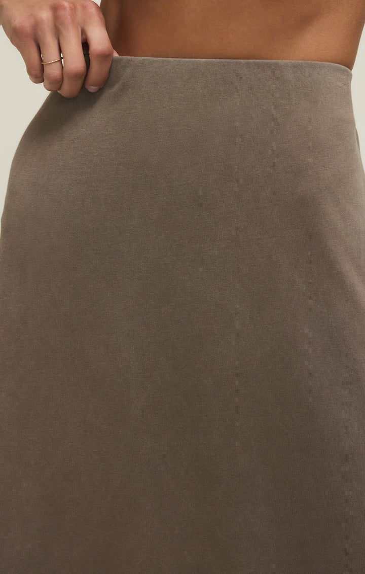A close-up shows a hand holding the elastic waistband of the Zertz GAIL MIDI SKIRT PERFECTLY TAUPE, a high-waisted cotton jersey skirt. The person wears a thin ring, set against a neutral, light background.
