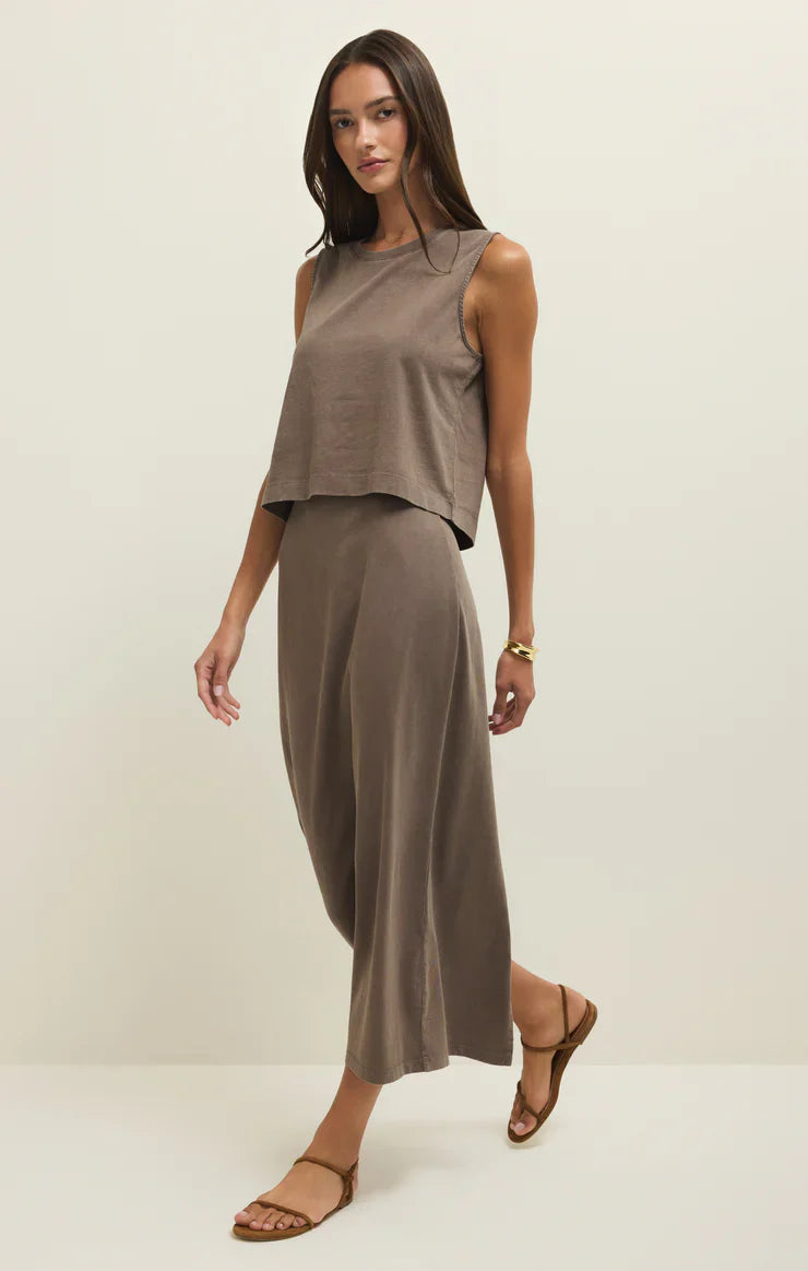 A woman in the Zertz GAIL MIDI SKIRT PERFECTLY TAUPE, paired with a loose, sleeveless brown top and flat sandals, stands against a plain light background, looking ahead with one foot forward.