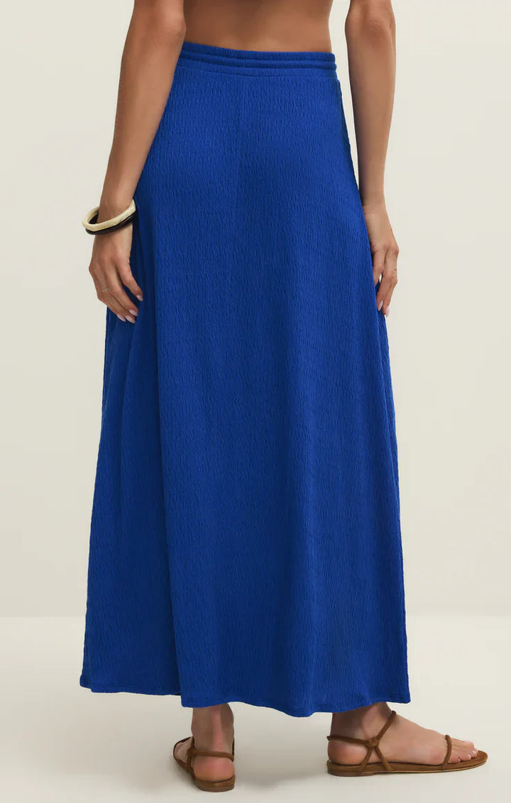 A woman stands with her back to the camera, wearing the bright blue Zertz IDRIS MAXI SKIRT with an elastic waistband, brown sandals, and a chunky white bracelet against a plain light background.