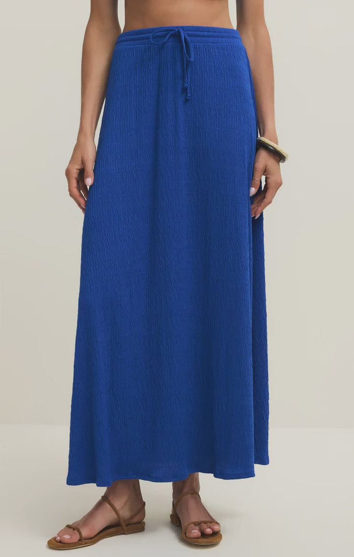 A person wearing the Zertz IDRIS MAXI SKIRT—a textured, bright blue pucker knit skirt with an elastic waistband—paired with brown flat sandals and a gold bracelet, stands against a plain light background.