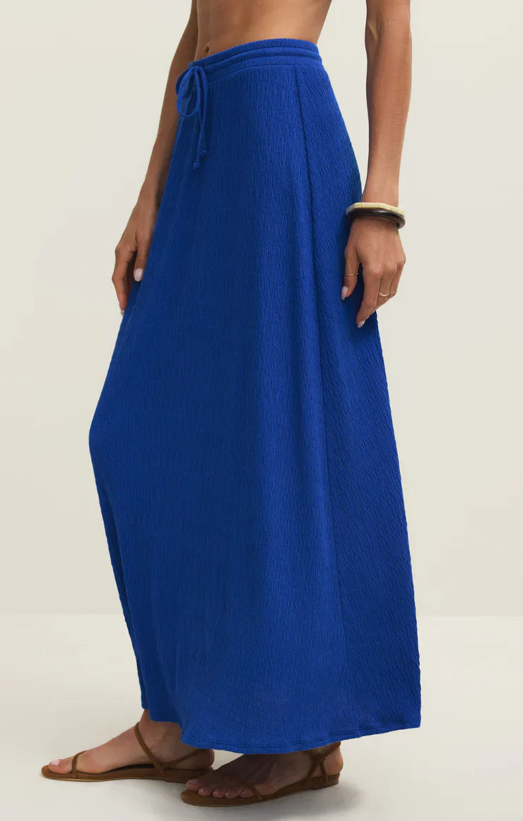 A person wears the Zertz IDRIS MAXI SKIRT in bright blue—an ankle-length pucker knit skirt with an elastic waistband and tie—paired with brown flat sandals and a chunky bracelet, set against a plain light beige background.