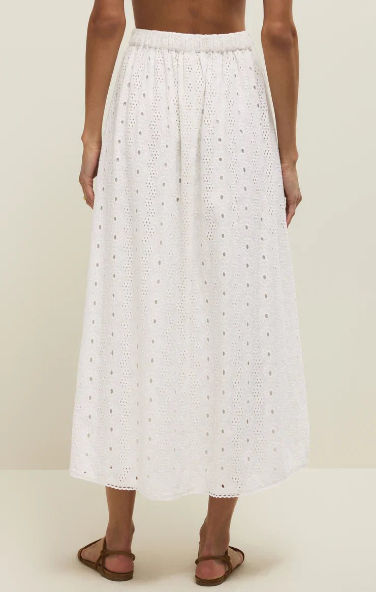 A person wears the Zertz BRIA EYELET SKIRT, a white cotton midi with an elastic waistband, flowy textured fabric, and delicate eyelet embroidery, standing barefoot against a neutral background.