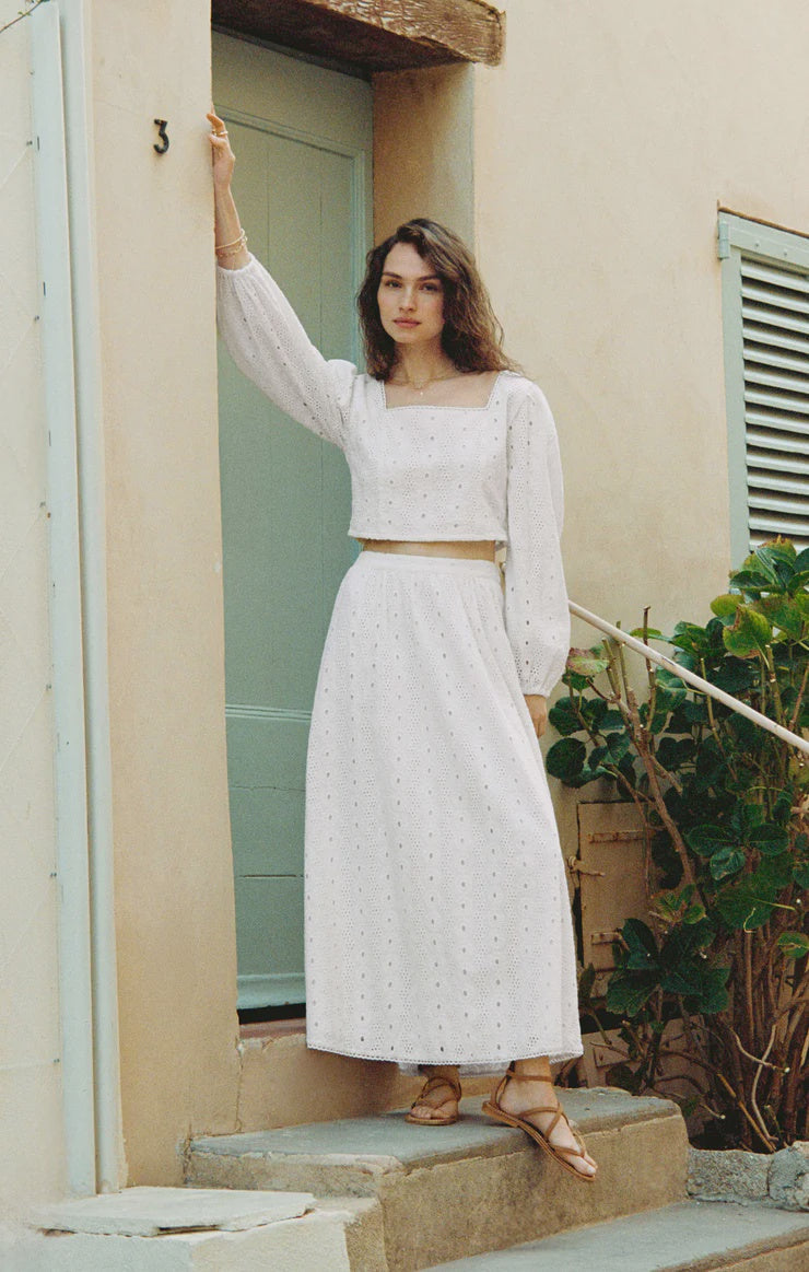 A woman stands on steps outside a pale blue door, wearing the Zertz BRIA EYELET SKIRT—a white, cotton midi skirt with an elastic waistband—paired with a matching long-sleeve crop top and sandals. She looks at the camera, one arm resting on the doorframe.