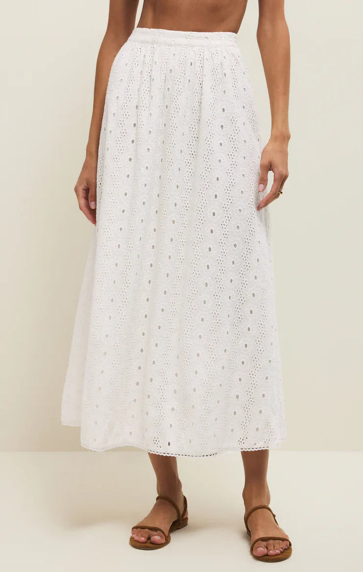 A woman wears the Zertz BRIA EYELET SKIRT, a white cotton midi with delicate eyelet detail and an elastic waistband, standing barefoot on a neutral background. Only her lower torso, skirt, and feet are visible.