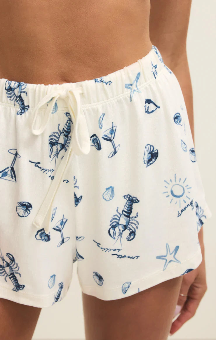 Close-up of a person wearing LOST AT SEA SHORT by Zertz—white jersey shorts with blue vacation print of lobsters, seashells, starfish, cocktails, and suns. Perfect loungewear for relaxing days.