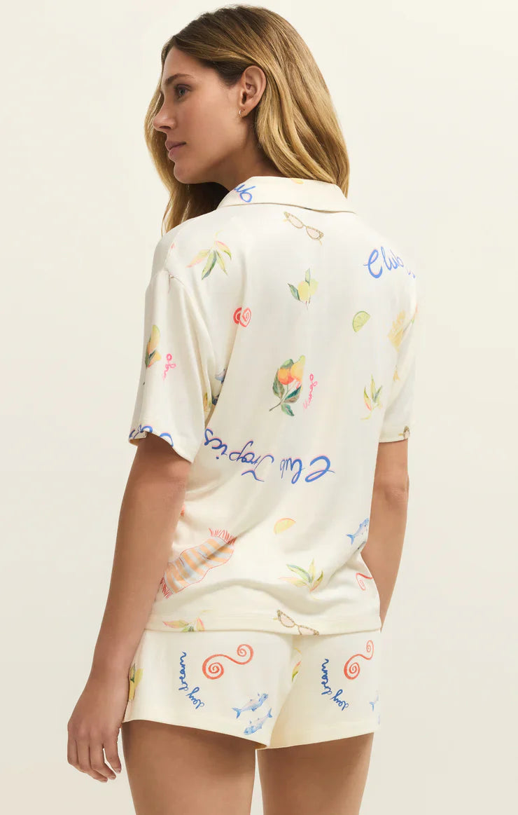 A woman with light brown hair models the Zertz BEACH DAYS PJ TOP, a relaxed-fit, cream pajama set in silky jersey with a colorful whimsical print, while standing facing away from the camera against a plain backdrop.