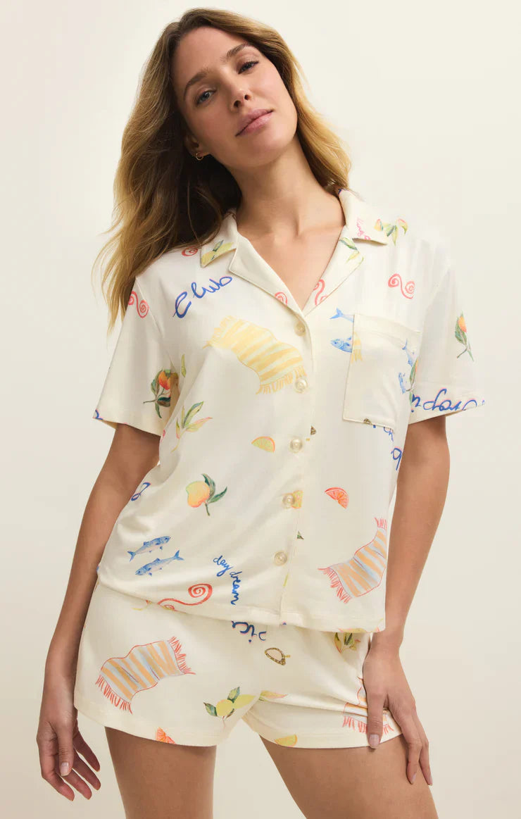 A woman models the Zertz BEACH DAYS PJ TOP, a relaxed-fit silky jersey short-sleeve lounge top with seashell and fruit print, paired with matching shorts, against a plain light background.