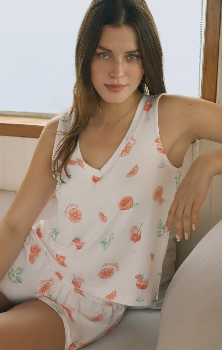 A young woman with long brown hair sits on a couch near a window, wearing the Zertz SPRITZ TANK—a lightweight v-neck pajama set adorned with colorful oranges, leaves, and drinks. She looks at the camera with a slight smile.