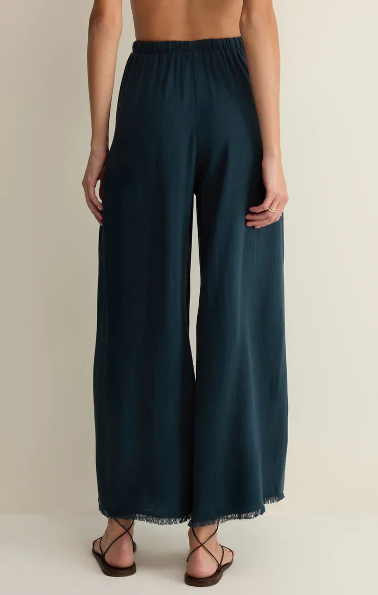 A person faces away, wearing Zertz SOREN PANT—high-rise linen pants with a frayed hem and elastic waistband—paired with flat black sandals against a plain, neutral background.