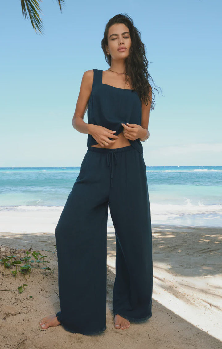 A woman stands barefoot on a sandy beach in a sleeveless navy blue top and Zertz SOREN PANT—high-rise linen pants with an elastic waistband—against the backdrop of the ocean and blue sky.