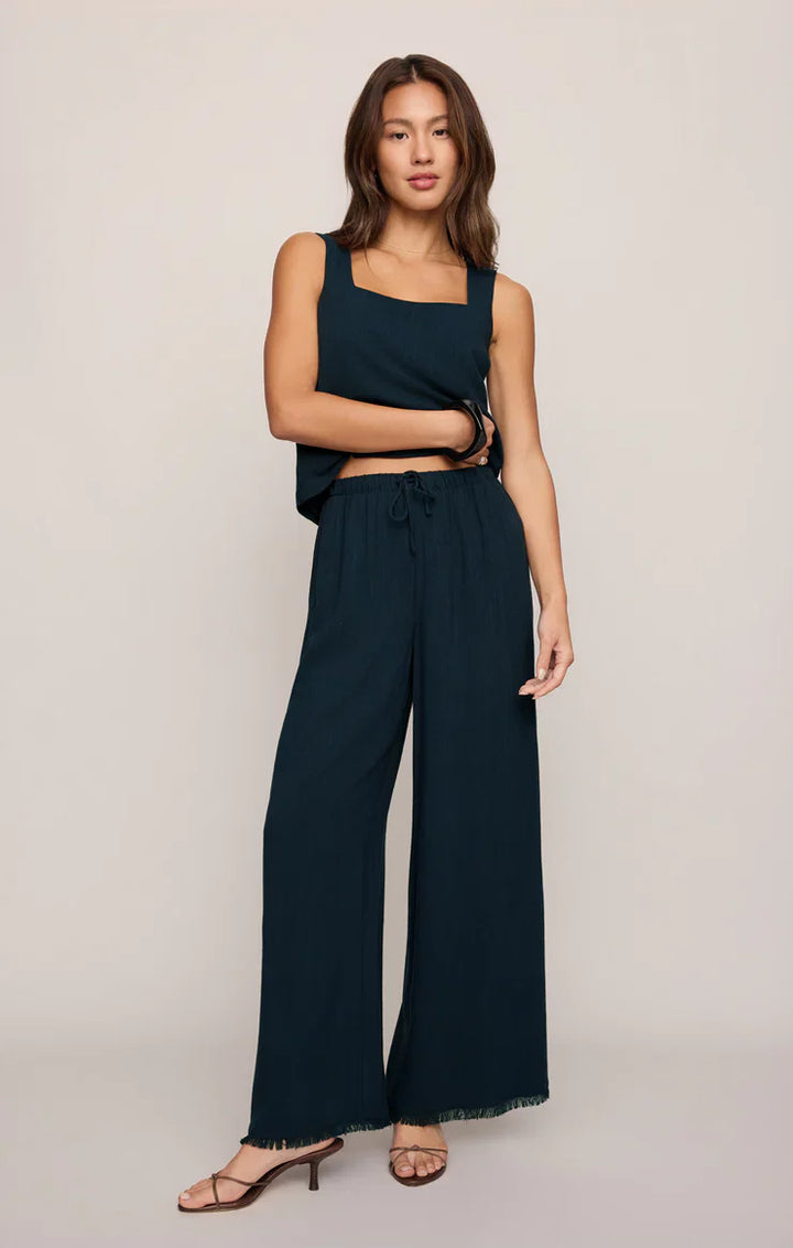 A woman with long hair and minimal accessories models Zertz’s SOREN PANT—a dark teal, high-rise linen pant with an elastic waistband—paired with a matching sleeveless square-neck top and heeled sandals against a plain background.
