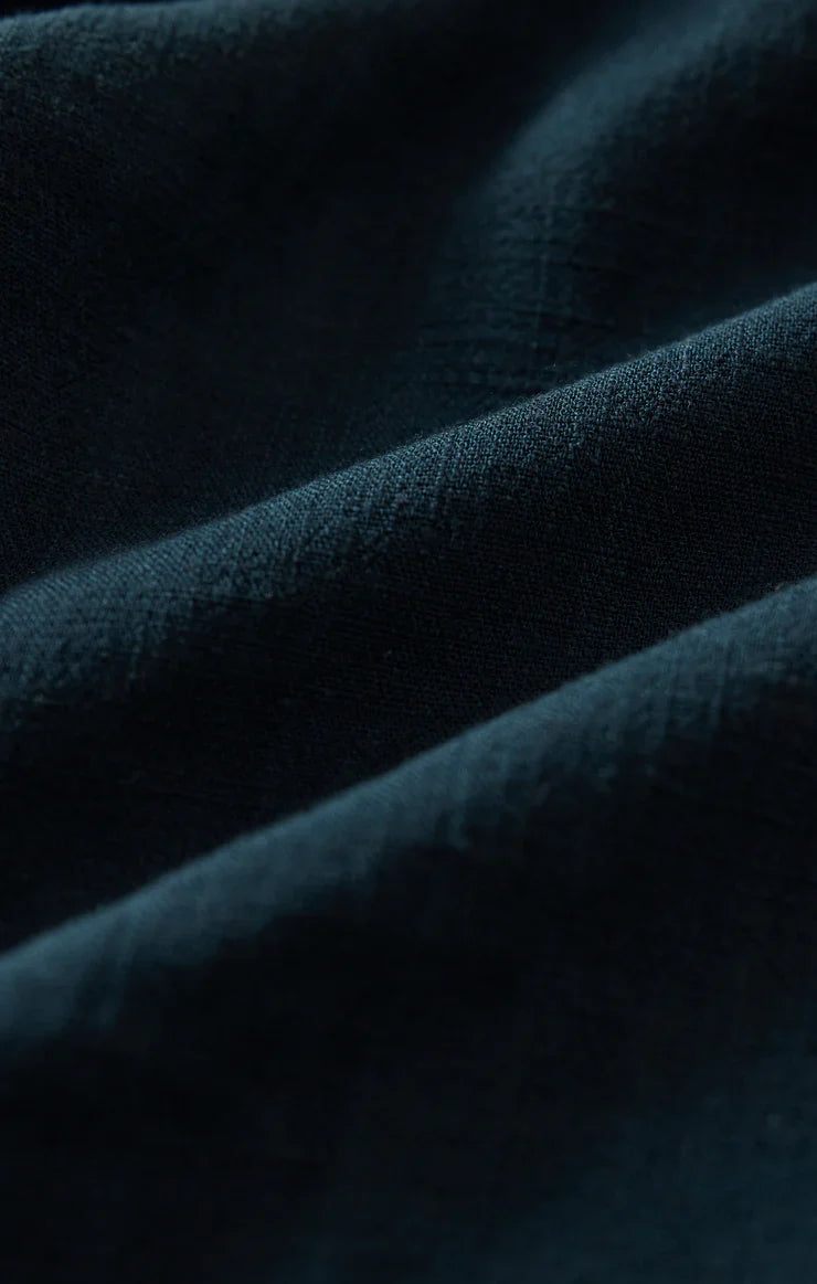Close-up of the dark green, textured fabric from Zertz’s SOREN PANT reveals fine lines and gentle folds in the high-rise linen, casting soft shadows across its surface.