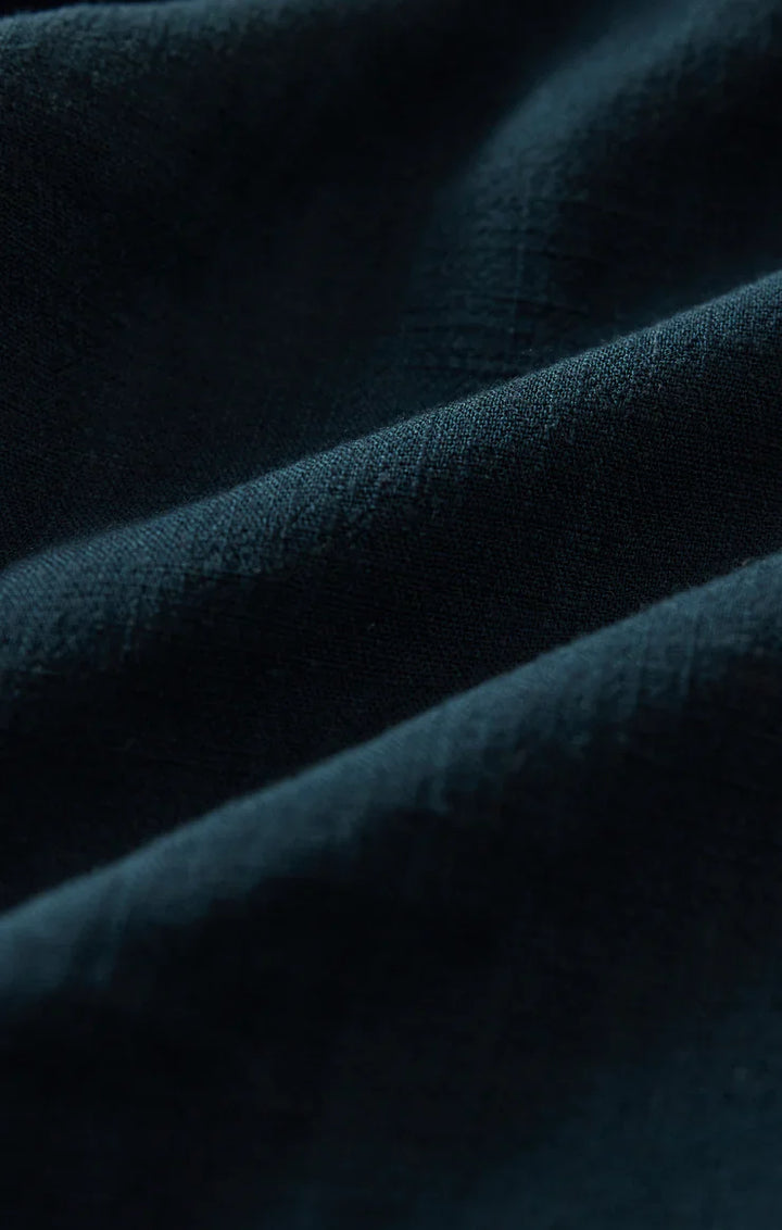 Close-up of the dark green, textured fabric from Zertz’s SOREN PANT reveals fine lines and gentle folds in the high-rise linen, casting soft shadows across its surface.