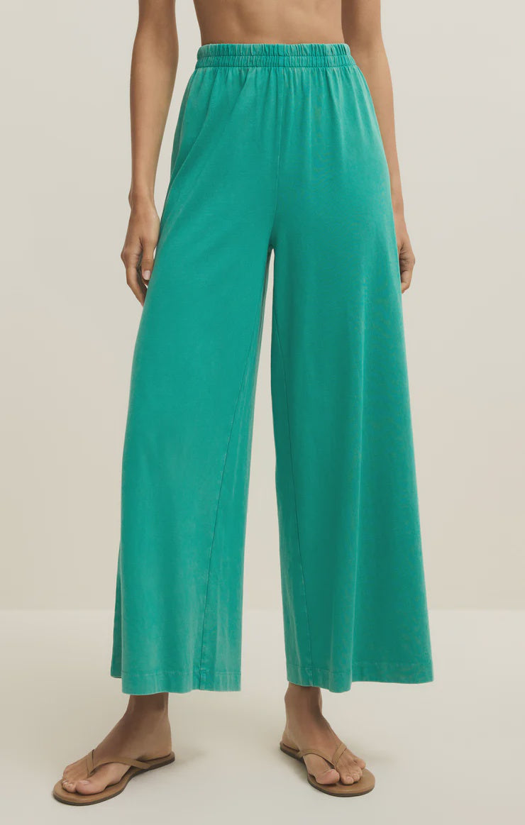 A person stands against a plain background wearing Zertz SCOUT JERSEY CROP FLARE PANT in teal, featuring a loose fit, high waist, wide flare legs, and elastic waistband, paired with tan flip-flops.