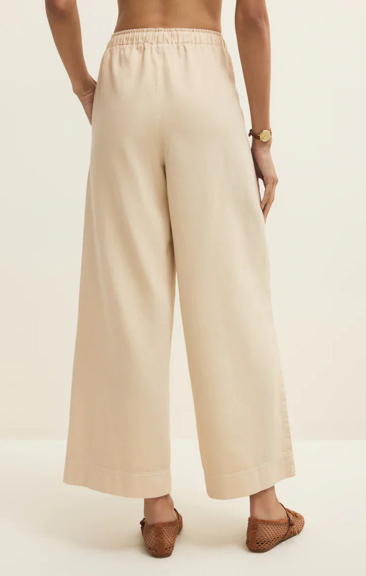 A person is seen from the back wearing Zertz DAYCATION PANT—high-waisted, wide leg with an elastic waistband—paired with brown woven flats and a watch, set against a plain light background.