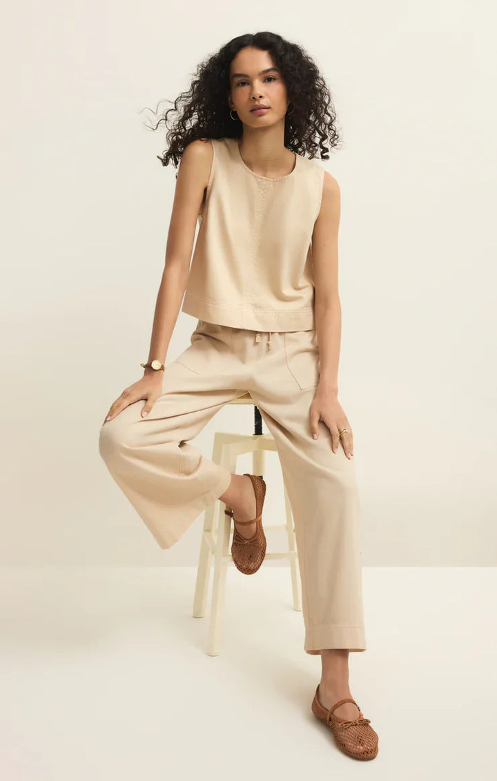 A person with curly hair sits on a white stool, wearing a sleeveless beige top, Zertz DAYCATION PANT with an elastic waistband, and brown woven slip-on shoes. They accessorize with a watch and rings against a plain light background.