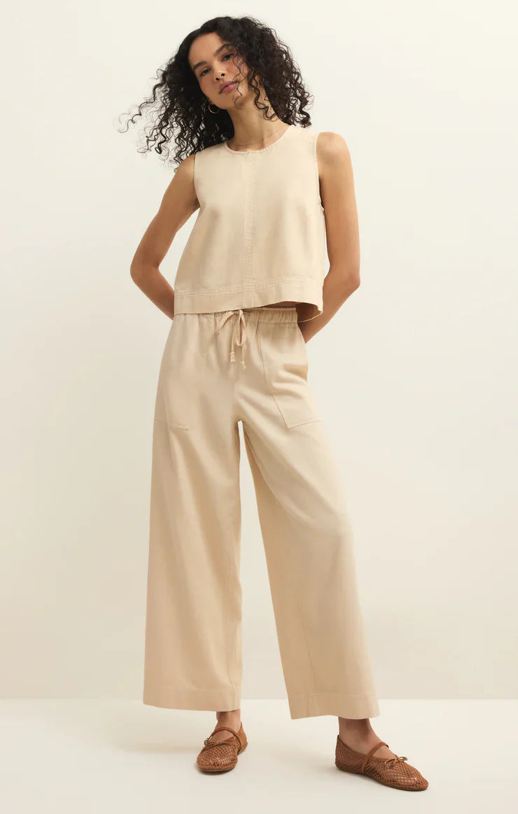 A woman with curly hair wears the Zertz DAYCATION PANT—wide-leg, beige, with an elastic waistband—paired with a sleeveless loose top and woven brown flats, standing against a plain light background.