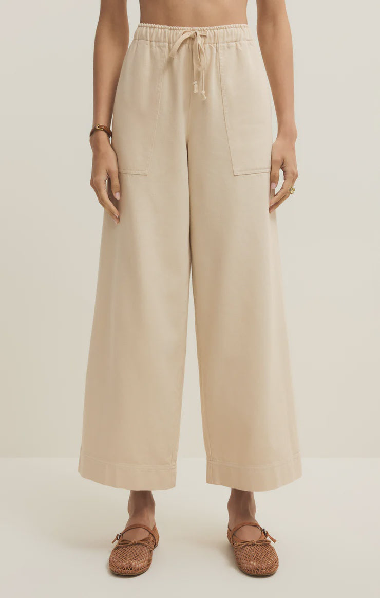 A person wears the Zertz DAYCATION PANT—high-waisted, wide-leg beige twill pants with front patch pockets and an elastic drawstring waist—paired with brown woven flats, standing against a plain light background.