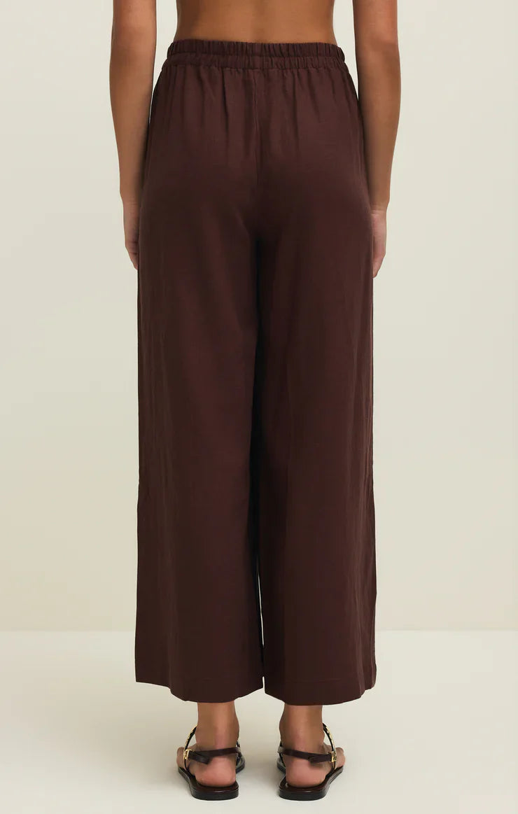 A person wears the Zertz SCOUT LINEN PANT—loose-fitting, high-rise, wide leg brown linen-blend pants with an elastic waistband—paired with black sandals. A light background highlights the relaxed silhouette.