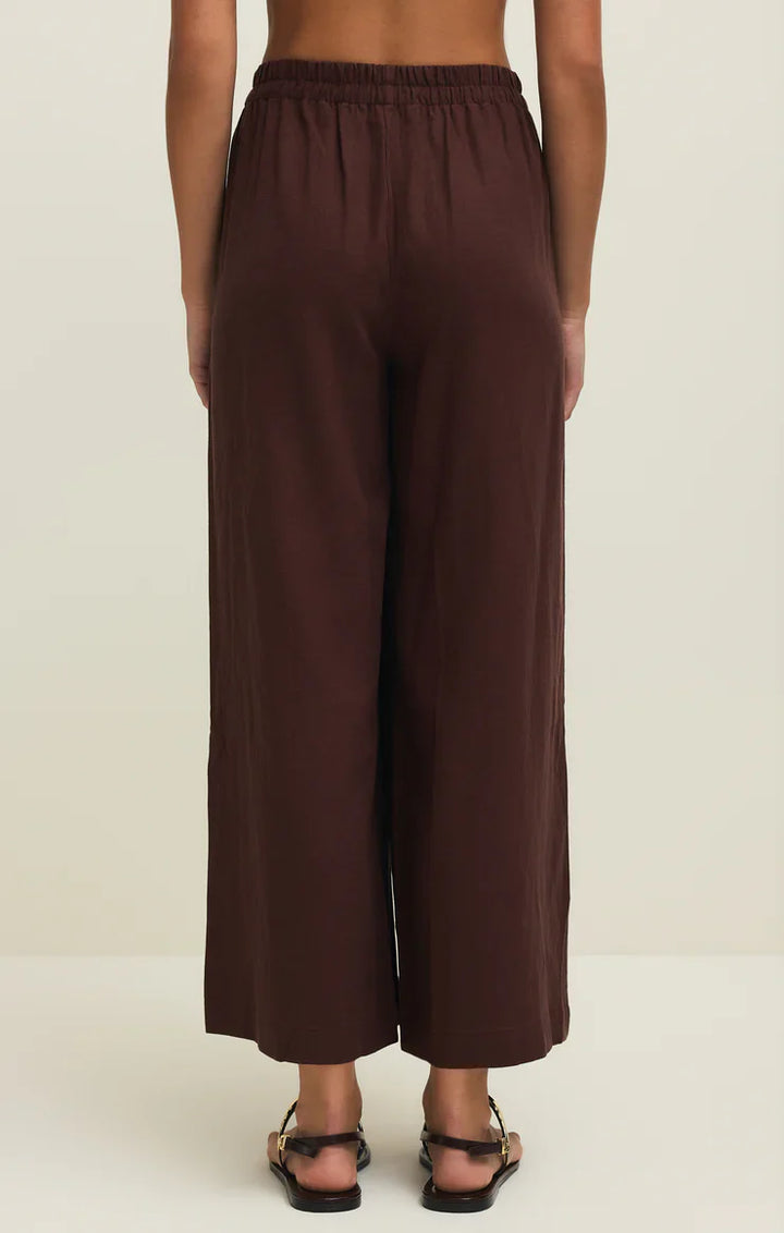 A person wears the Zertz SCOUT LINEN PANT—loose-fitting, high-rise, wide leg brown linen-blend pants with an elastic waistband—paired with black sandals. A light background highlights the relaxed silhouette.