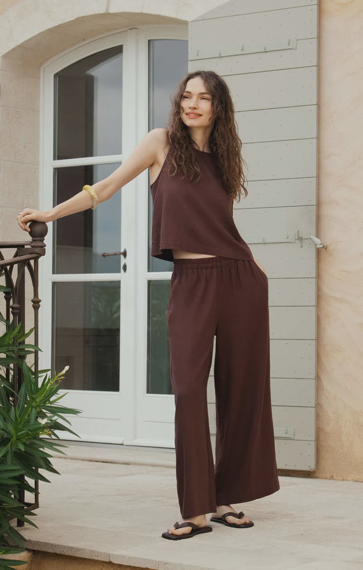 A woman with long wavy hair smiles outside by a railing, wearing Zertz's SCOUT LINEN PANT and a matching sleeveless brown top, paired with black sandals and a gold bangle. Behind her is a large white door and light-colored wall.