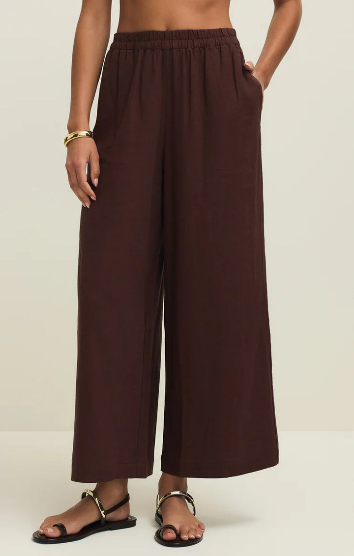 A person wears the Zertz SCOUT LINEN PANT—loose, high rise, wide leg brown linen blend—with hands in pockets, paired with a gold bracelet and black sandals against a plain light background.
