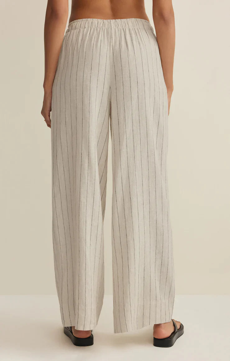 A person faces away wearing Zertz HENRI STRIPED WIDE LEG PANT—high-rise linen pants with an elastic waistband and thin vertical black stripes—paired with black slide sandals, set against a neutral background.