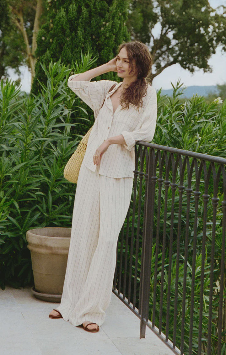 A woman stands on a balcony by green plants, smiling with her hand in her hair and holding a yellow woven bag. She is wearing the Zertz HENRI STRIPED WIDE LEG PANT, featuring loose linen fabric and an elastic waistband.
