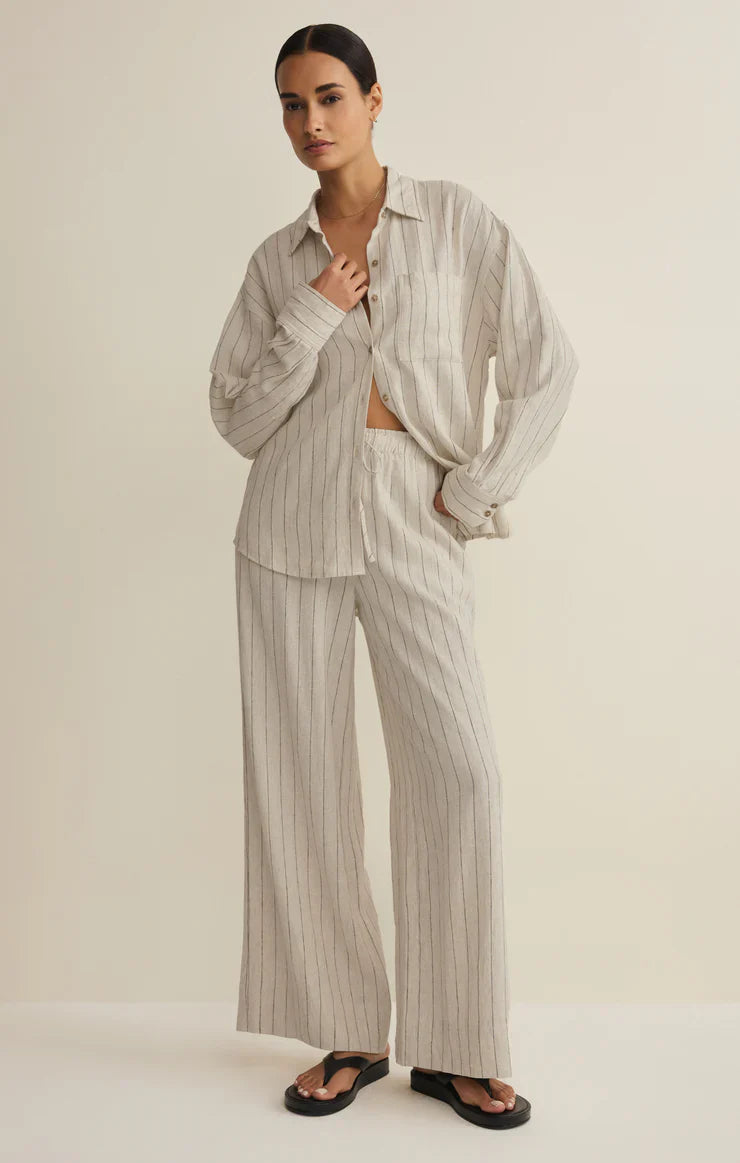 A woman poses against a plain background wearing Zertz’s HENRI STRIPED WIDE LEG PANT in beige, styled with a loose, matching pinstriped shirt and black sandals. She has one hand in her pocket.