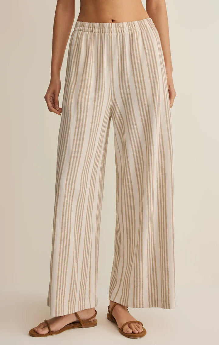 A person wears the Zertz SCOUT KNIT STRIPED PANT, featuring high-waisted, wide legs with beige and white vertical stripes and a comfy pull-on waistband, paired with brown flat sandals against a neutral backdrop.