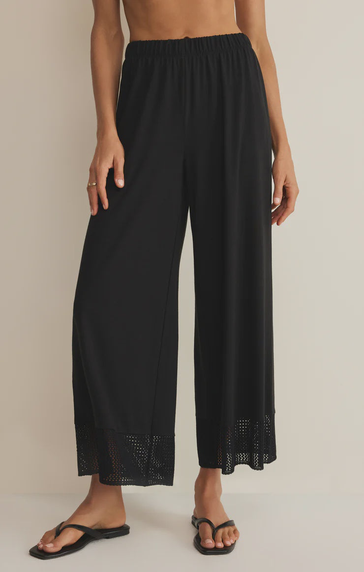 A person wears the Zertz BANYAN PANT—black, relaxed straight-leg with an elastic waistband and eyelet lace trim at the hems—paired with black flip-flops, standing against a plain light-colored background.