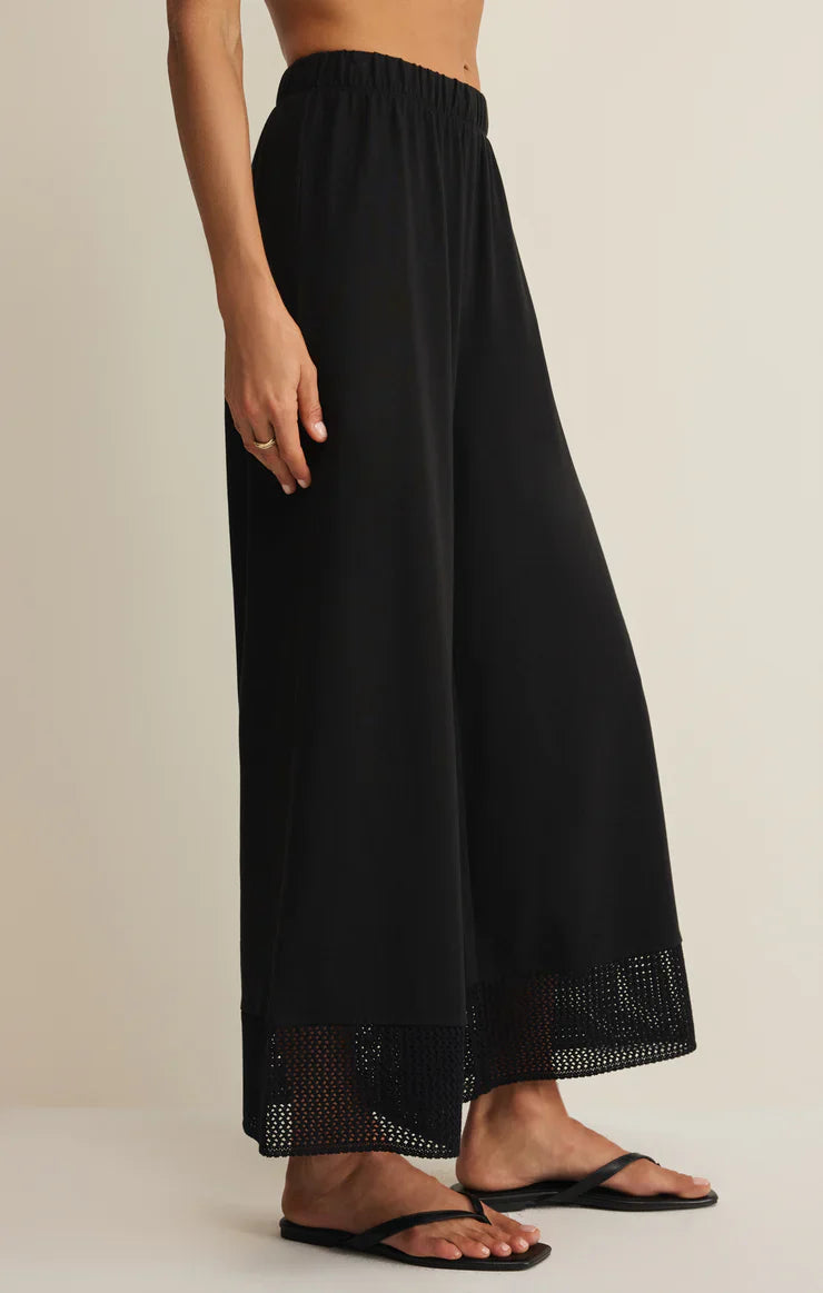 A person stands against a plain light background wearing Zertz’s BANYAN PANT—high-rise, relaxed straight-leg black pants with mesh hem detailing—paired with black flip-flops.