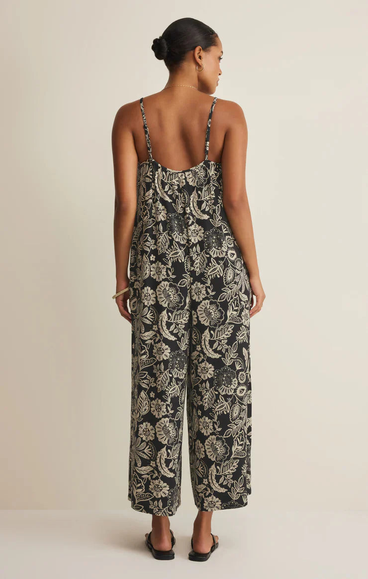 A woman with a low bun wears the Zertz DAY OFF LUZ FLORAL JUMPSUIT, a black, relaxed-fit style with wide legs. She completes her look with flat sandals and stands against an off-white background.