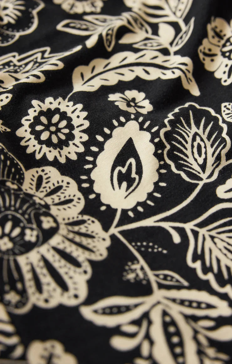 Close-up of the DAY OFF LUZ FLORAL JUMPSUIT by Zertz: black stretch jersey fabric with off-white stylized floral patterns in a bold, decorative design. Slightly textured and creased, ideal for a relaxed fit.
