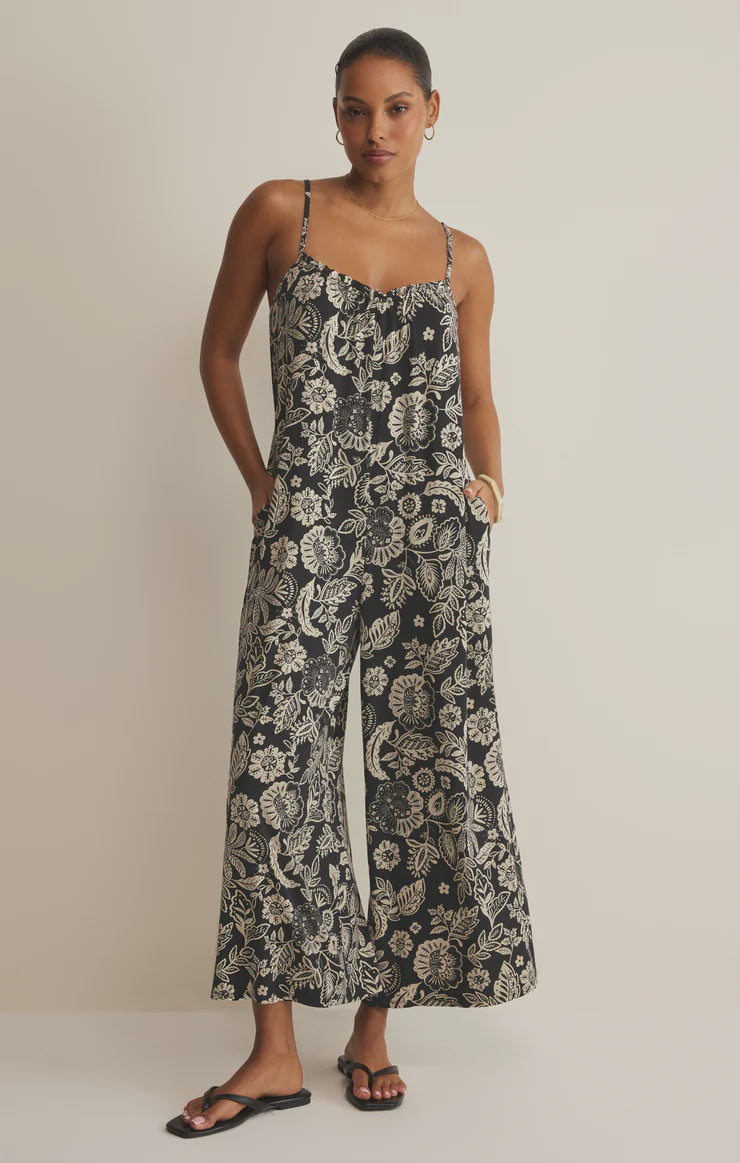 A woman wears the Zertz DAY OFF LUZ FLORAL JUMPSUIT, a relaxed fit, sleeveless stretch jersey with white florals and wide-leg cropped pants. She pairs it with black sandals, stands against a plain background with one hand in her pocket and a neutral expression.