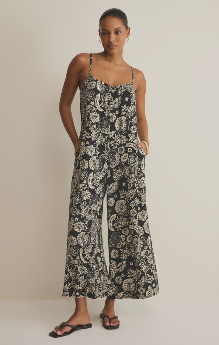 A woman wears the Zertz DAY OFF LUZ FLORAL JUMPSUIT, a relaxed fit, sleeveless stretch jersey with white florals and wide-leg cropped pants. She pairs it with black sandals, stands against a plain background with one hand in her pocket and a neutral expression.