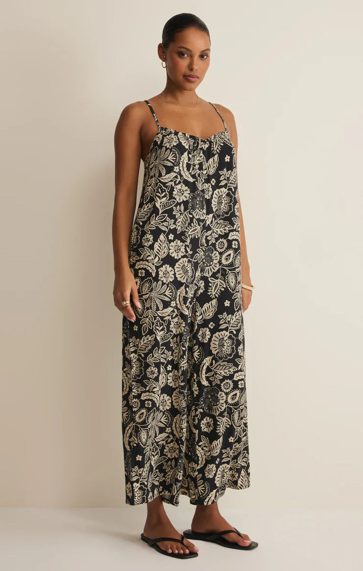 A woman stands against a plain light wall, wearing the Zertz DAY OFF LUZ FLORAL JUMPSUIT—an ankle-length, relaxed fit black jumpsuit with an intricate beige floral pattern—paired with black sandals and gold hoops. Hair is styled in a neat bun.