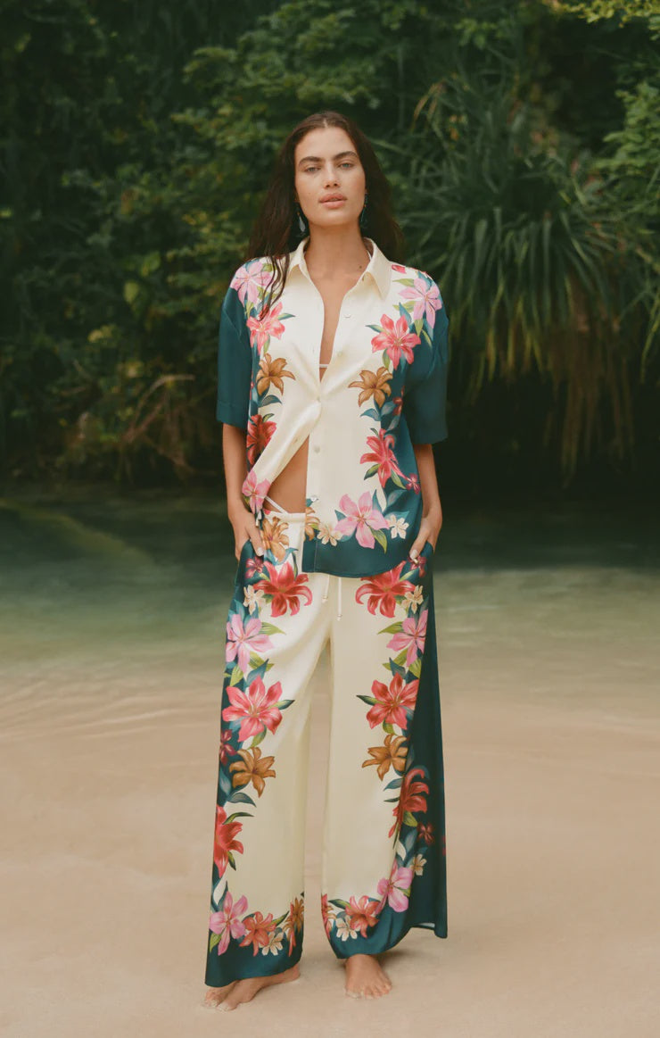 A woman stands barefoot on a sandy beach, framed by lush green foliage, wearing Zertz’s LANI KIWI PANT—vacation-ready wide leg satin pants—and a pajama-style top with a vibrant pink and orange tropical hibiscus print.