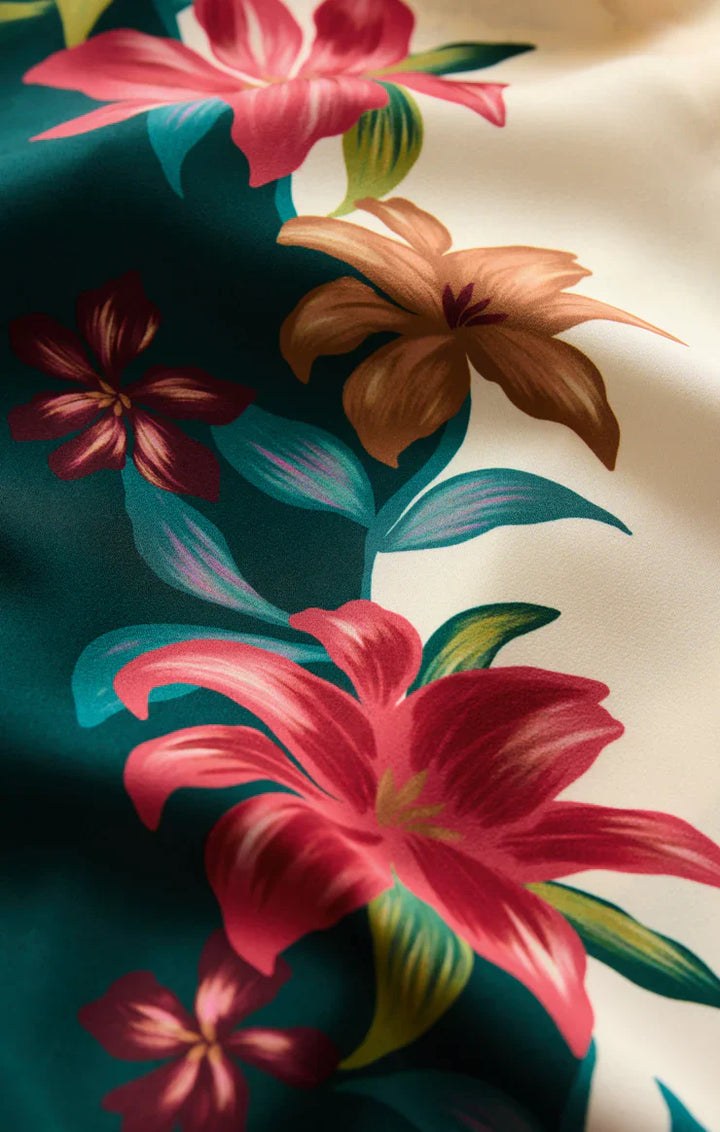 A close-up of the LANI KIWI PANT by Zertz shows a vibrant tropical hibiscus print with bold pink, red, and brown flowers plus green leaves on cream and dark green satin—perfect for vacation-ready wide leg style.
