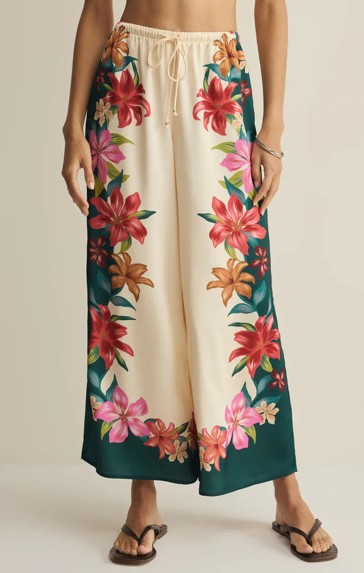 A person in Zertz LANI KIWI PANT—vacation-ready wide-leg cream pants with a drawstring waist and vibrant tropical hibiscus print down the sides and bottom—paired with brown flip-flops.