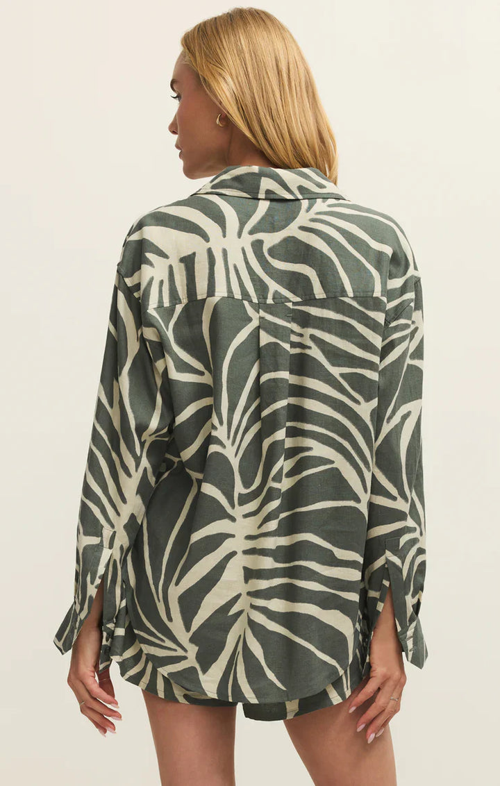 A woman with blonde hair wears the Zertz ALDI TOVA PALM BUTTON UP TEA LEAF, an oversized long-sleeved shirt with a bold green and white palm print, standing with her back to the camera against a plain background.