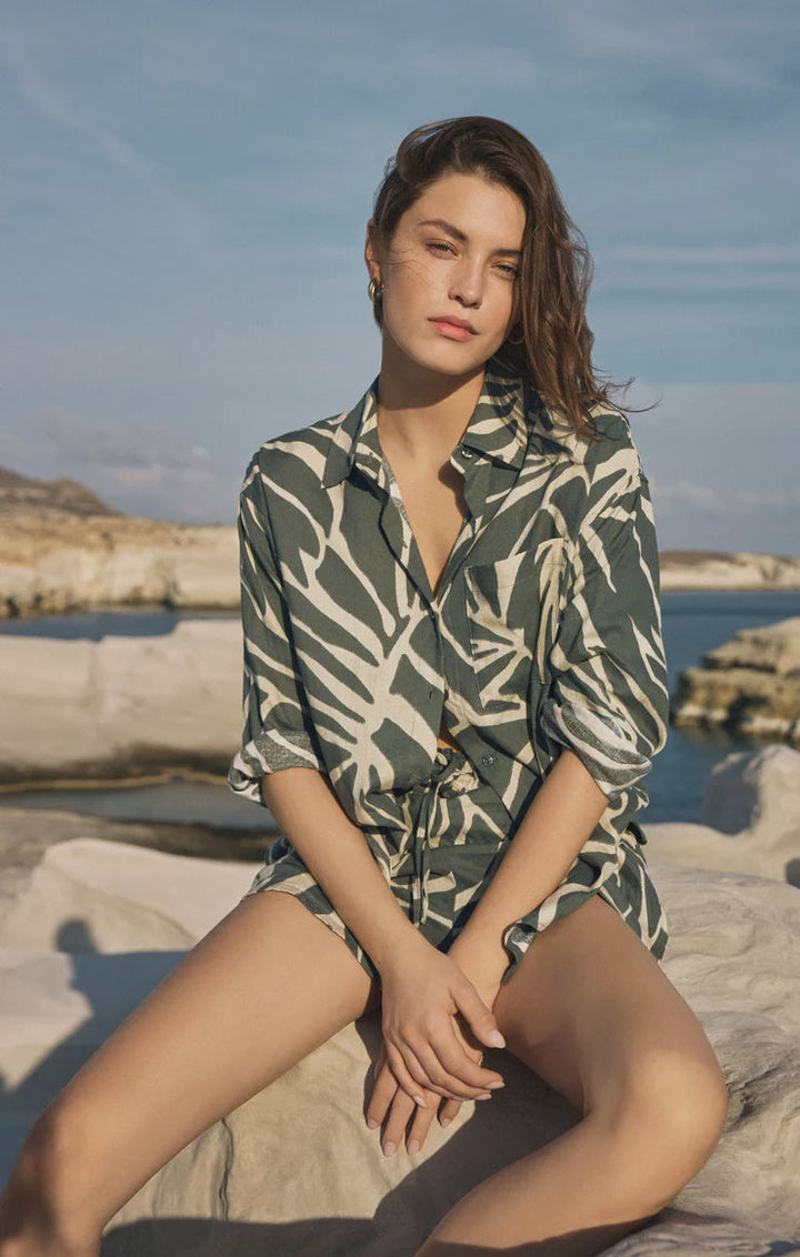 A person with wavy brown hair sits on white rocks by the water, wearing the Zertz ALDI TOVA PALM BUTTON UP TEA LEAF top, looking at the camera with a relaxed expression under a blue sky.