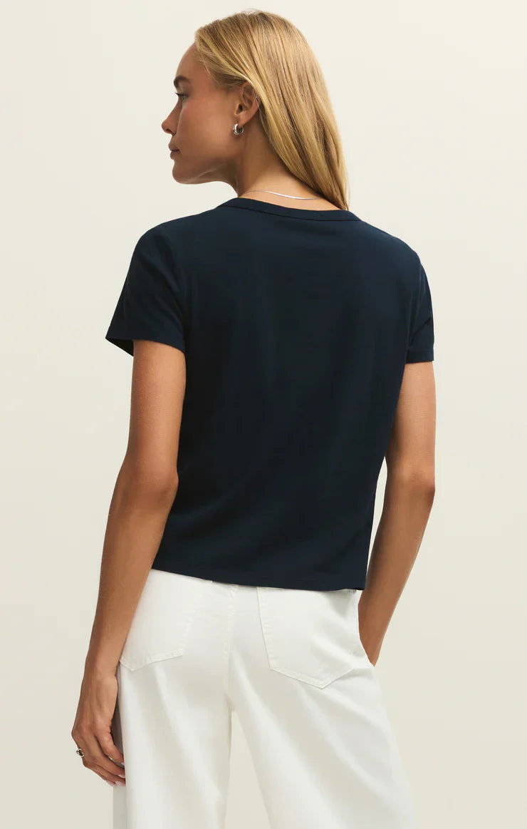 A woman with straight blonde hair is seen from the back, wearing the relaxed-fit Zertz JUNIE TEE SHIRT in black and high-waisted white pants, standing against a plain light background.