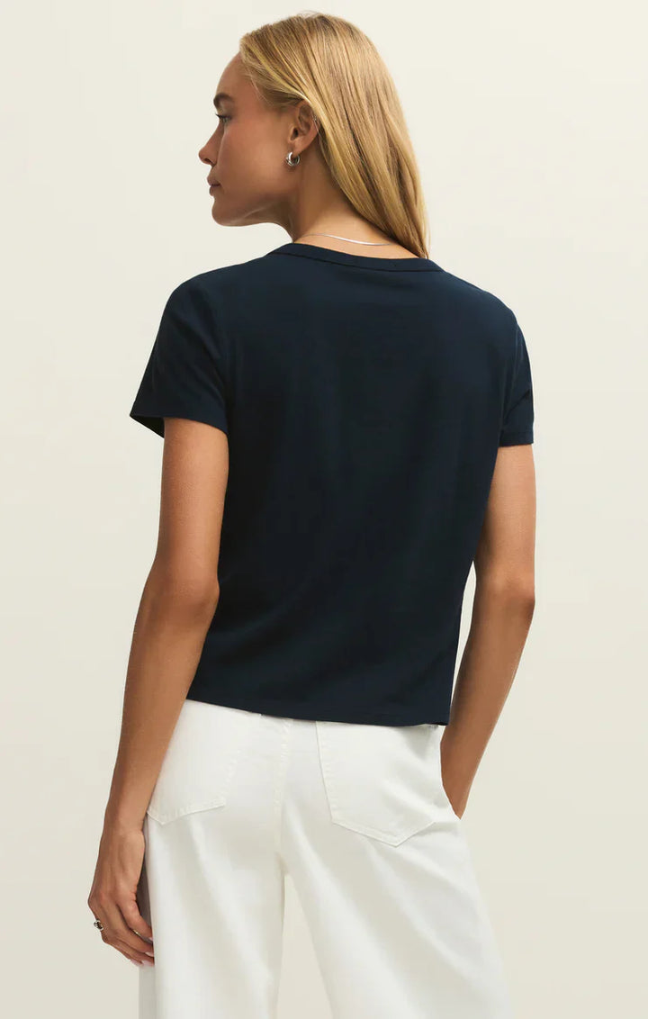 A woman with straight blonde hair is seen from the back, wearing the relaxed-fit Zertz JUNIE TEE SHIRT in black and high-waisted white pants, standing against a plain light background.