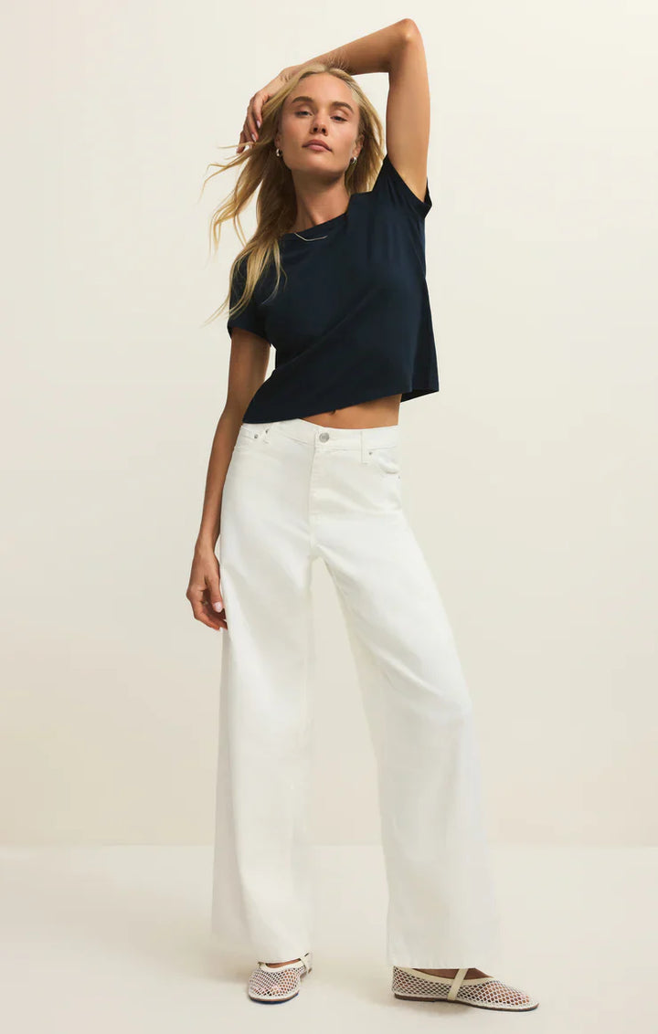 A woman poses against a plain background wearing the Zertz JUNIE TEE SHIRT, a navy blue 100% cotton crewneck tee, paired with high-waisted wide-leg white pants and patterned flats. She rests one hand on her head for a relaxed vibe.